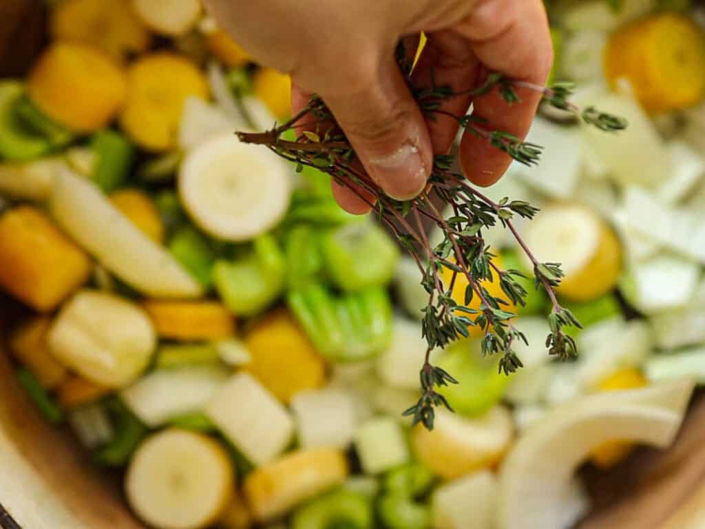 A hand holding fresh thyme sprigs over a mix of chopped yellow carrots, celery, onions, and other vegetables in a bowl.